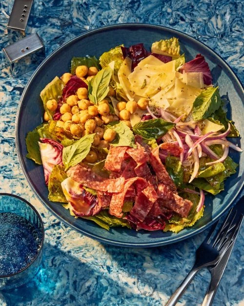 Overhead view of a vibrant salad featuring salami, lettuce, and chickpeas in a blue bowl set against a marble surface with flatware.