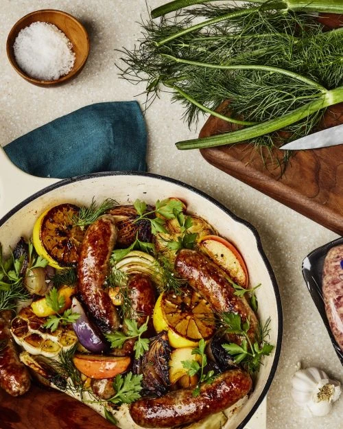 Overhead shot of skillet with cooked sausages, sliced lemons, and fresh herbs alongside salt, cutting board, and kitchen towel.