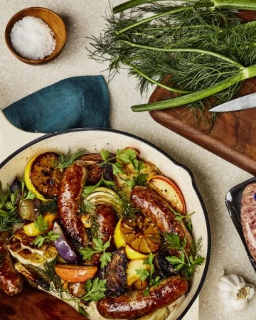 Overhead shot of skillet with cooked sausages, sliced lemons, and fresh herbs alongside salt, cutting board, and kitchen towel.