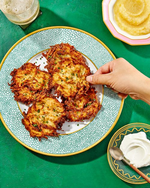 Overhead shot of golden latkes on a patterned plate, accompanied by applesauce, sour cream, and beer on a green surface.