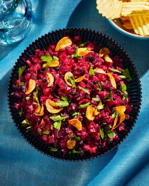 Overhead shot of vibrant beetroot salad in a black bowl, garnished with garlic and parsley, set on a blue tablecloth beside chips.
