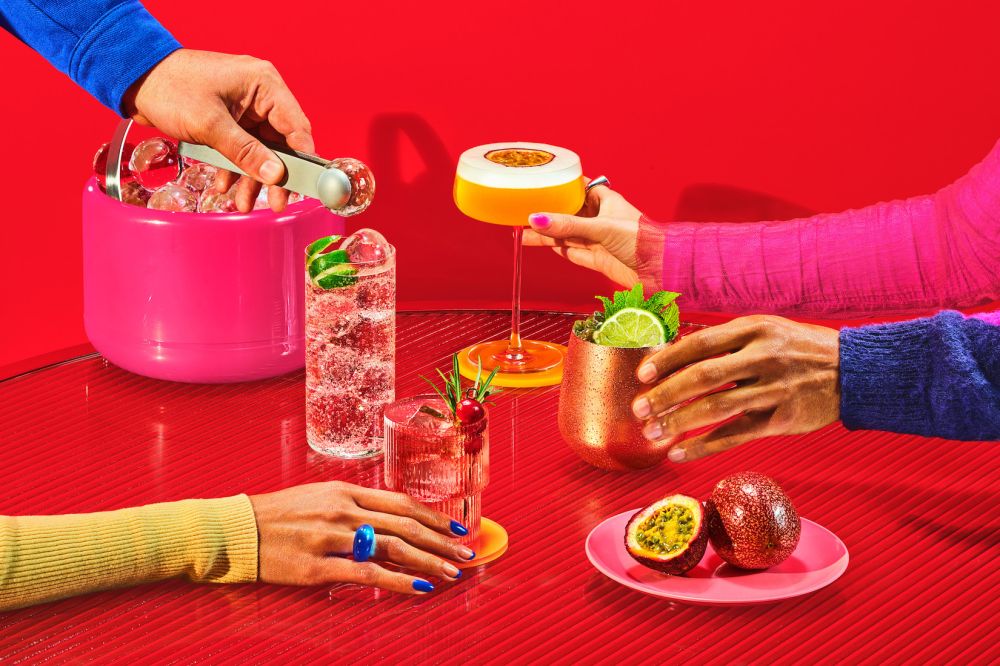 Several people around a red table, preparing and enjoying colorful tropical cocktails with ice and various garnishes and fruit.