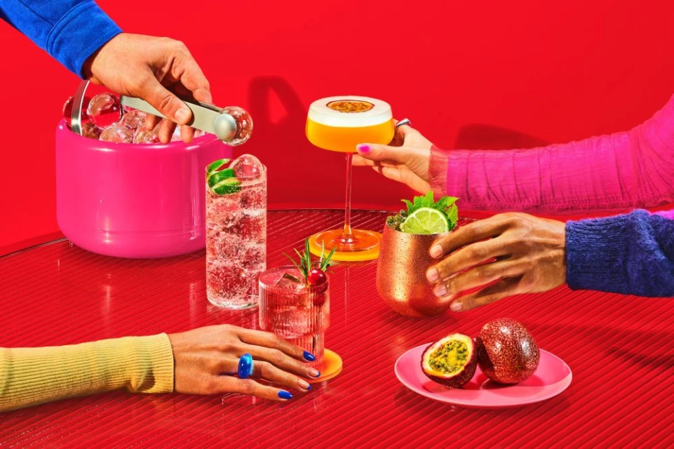 Several people around a red table, preparing and enjoying colorful tropical cocktails with ice and various garnishes and fruit.