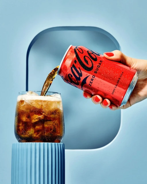 Coca-Cola Zero Sugar can pouring soda into a glass with ice, held by a hand with red nail polish, in front of a blue background.
