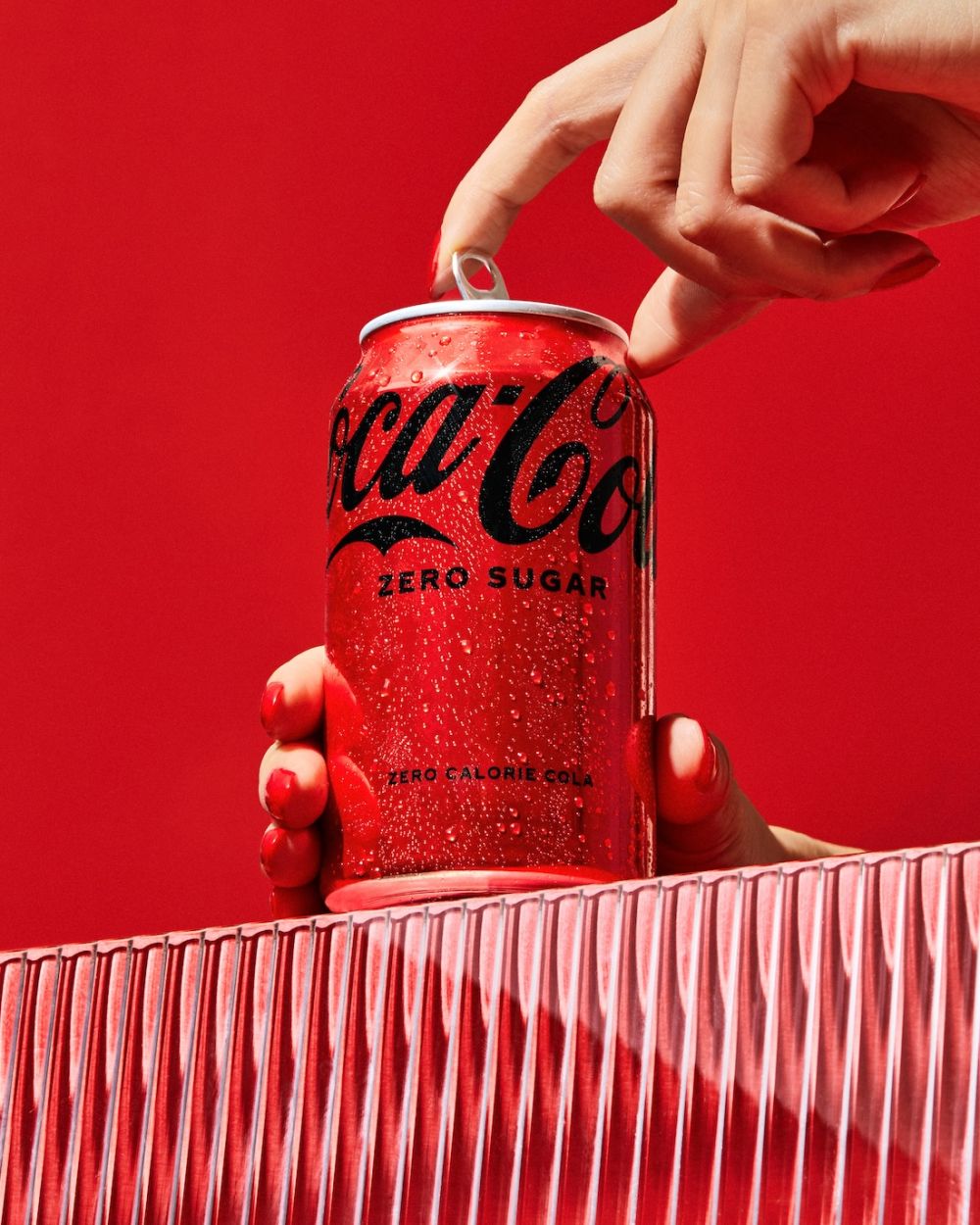 A red Coca-Cola Zero Sugar can is being held by a hand against a red background with textured glass surface beneath it.