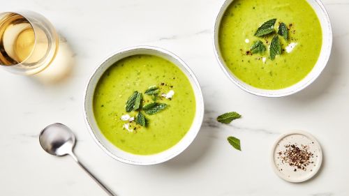 Overhead view of two bowls of vibrant green soup garnished with fresh mint, alongside a glass of white wine and spices on marble.