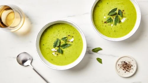 Overhead view of two bowls of vibrant green soup garnished with fresh mint, alongside a glass of white wine and spices on marble.