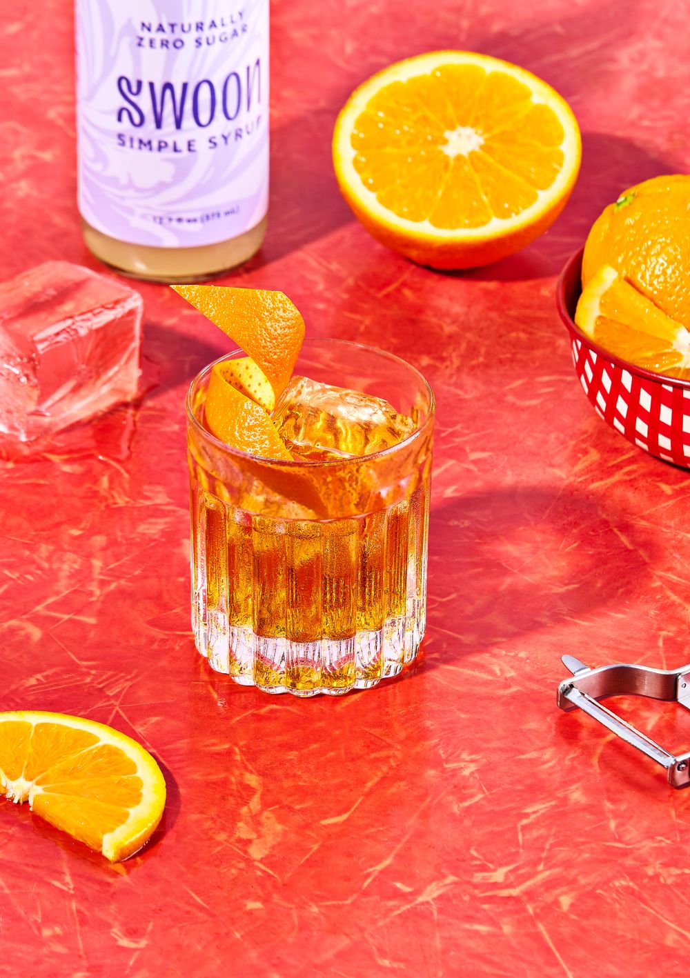 A cocktail in a glass with orange peel and ice sits on a red surface, accompanied by oranges and a Swoon simple syrup bottle.