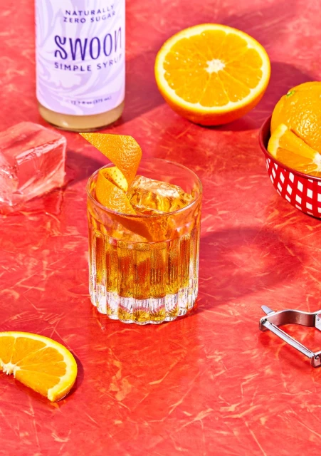 A cocktail in a glass with orange peel and ice sits on a red surface, accompanied by oranges and a Swoon simple syrup bottle.