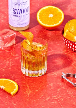 A cocktail in a glass with orange peel and ice sits on a red surface, accompanied by oranges and a Swoon simple syrup bottle.