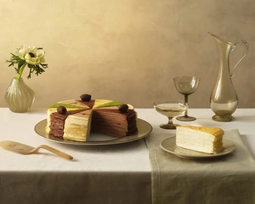 Still life shot of a mille crepe cake with slices missing, a glass of wine, and a vase of white anemones against a muted background.