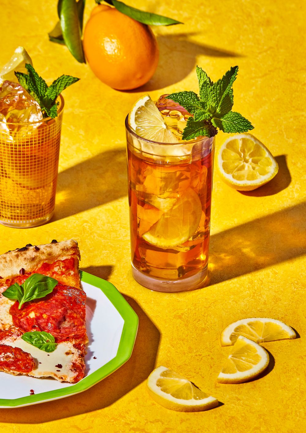 Overhead shot of a pizza slice on a plate next to cocktails, sliced lemons, and an orange on a yellow textured surface.