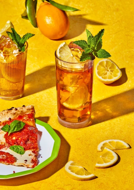 Overhead shot of a pizza slice on a plate next to cocktails, sliced lemons, and an orange on a yellow textured surface.
