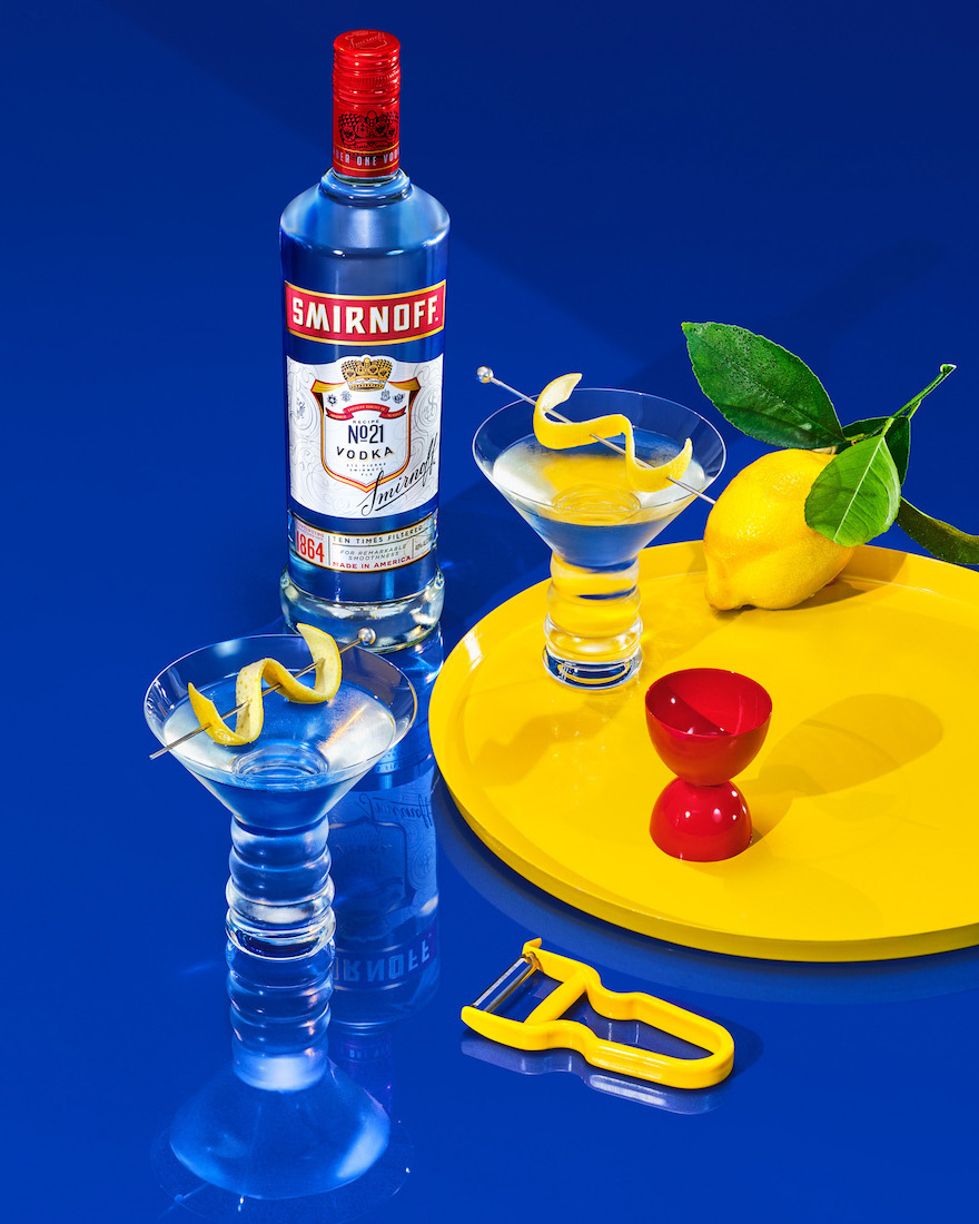 Smirnoff vodka bottle with martini glasses, lemon, and peeler on a blue background, showcasing barware and drink preparation.