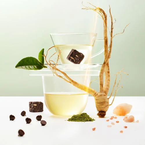 Still life studio shot showcases two clear glass cups filled with light liquid, chocolate pieces, ginseng root, tea leaf, and matcha powder.