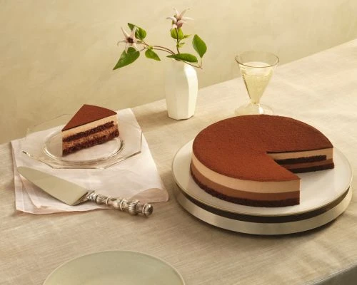 Still life of a three-layer chocolate cake with a slice removed, plated beside a cake server, drink, and a floral arrangement.