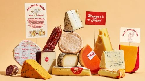 An assortment of cheeses and cured meats, including hard and soft cheeses, salami, and signage with food and promotional messaging.