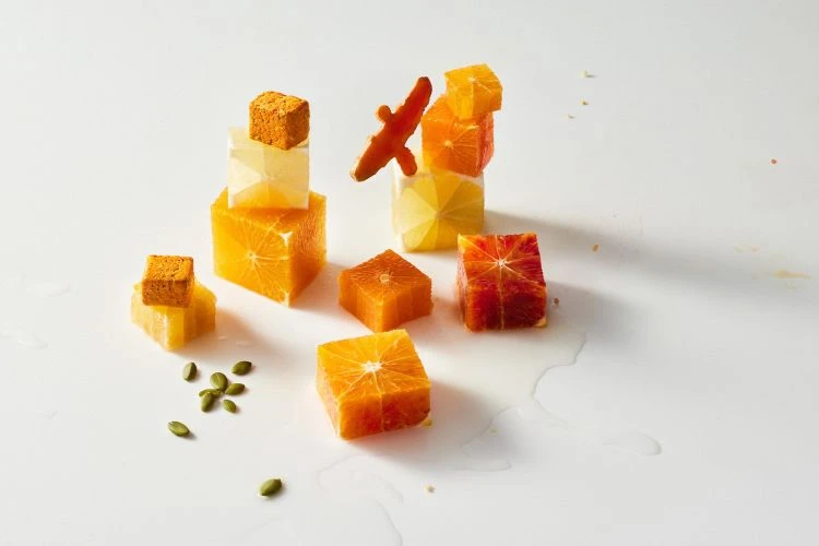 Stacked citrus cubes in vivid colors: orange, lemon, and blood orange on a bright white background, with scattered seeds and juice.