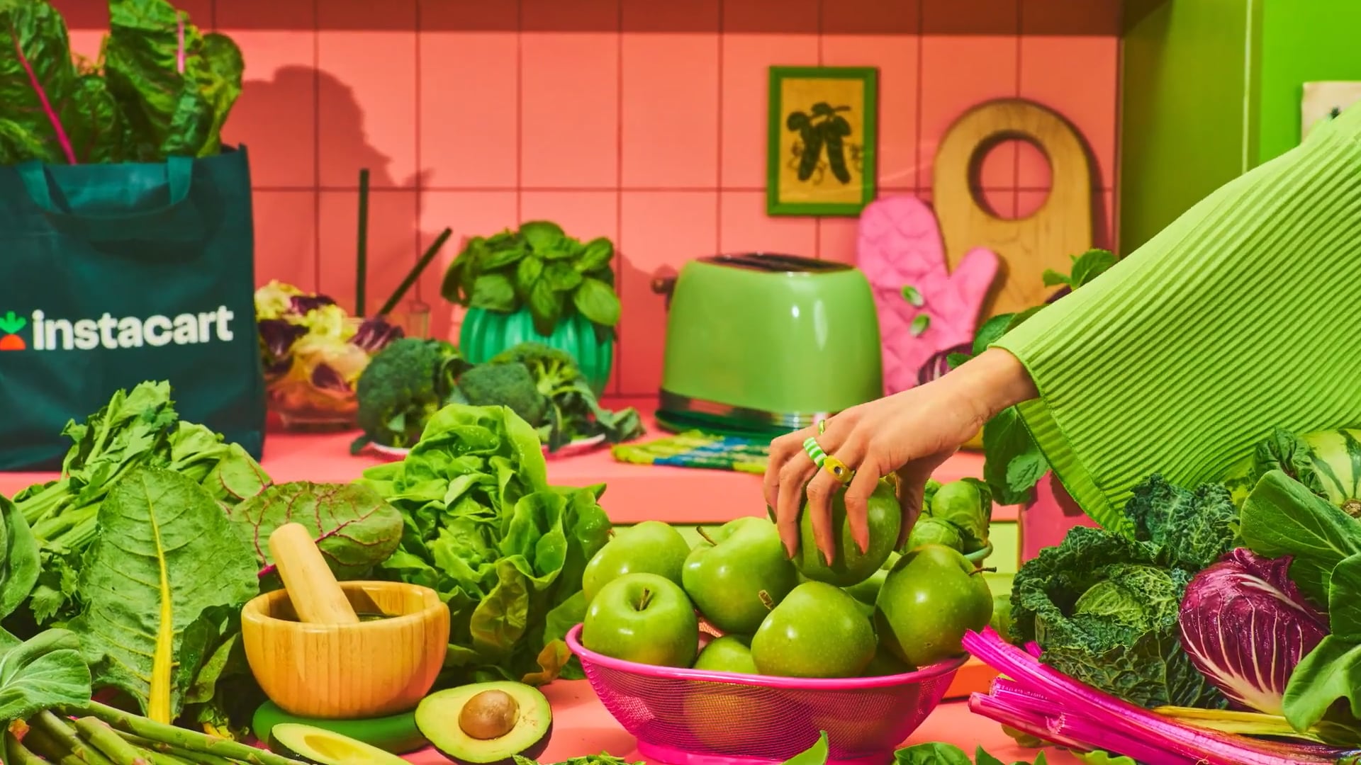 Fresh green apples in a pink wire basket surrounded by various vegetables and greens in a colorful kitchen scene with an Instacart bag.