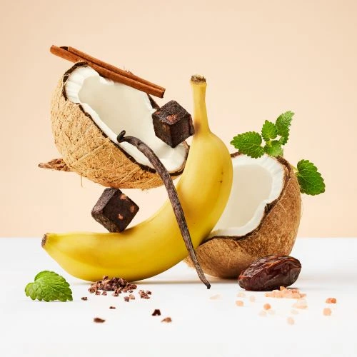 Close-up studio shot of a banana, coconut halves, and cinnamon sticks, with chocolate cubes and spices floating on a cream background.