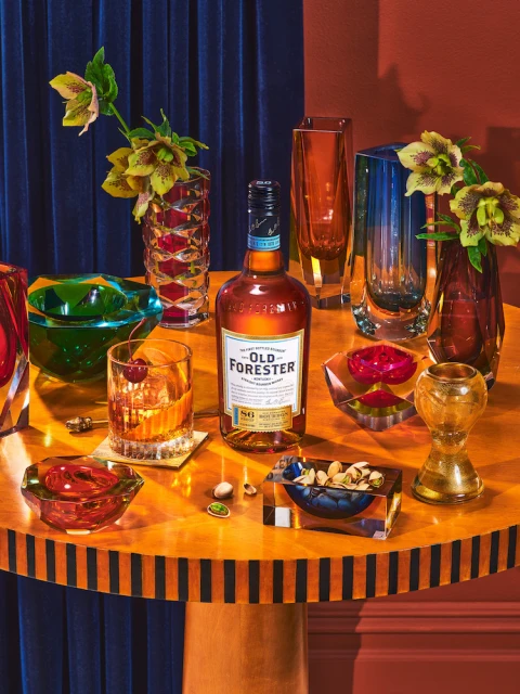 Old Forester bottle with glass of whiskey, cherries, various glassware on round wood table against a dark blue draped background.