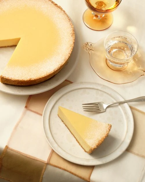 Overhead shot of a creamy cheesecake with a slice on a plate, accompanied by drinks, set against a light-colored background.