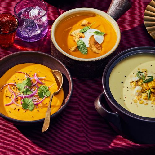 Overhead shot of three bowls of vibrant, colourful soups: orange butternut squash, creamy corn, served on a rich burgundy tablecloth.