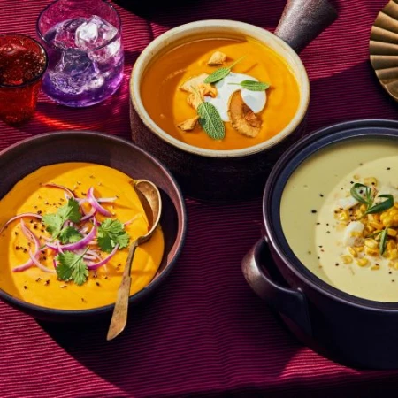 Overhead shot of three bowls of vibrant, colourful soups: orange butternut squash, creamy corn, served on a rich burgundy tablecloth.