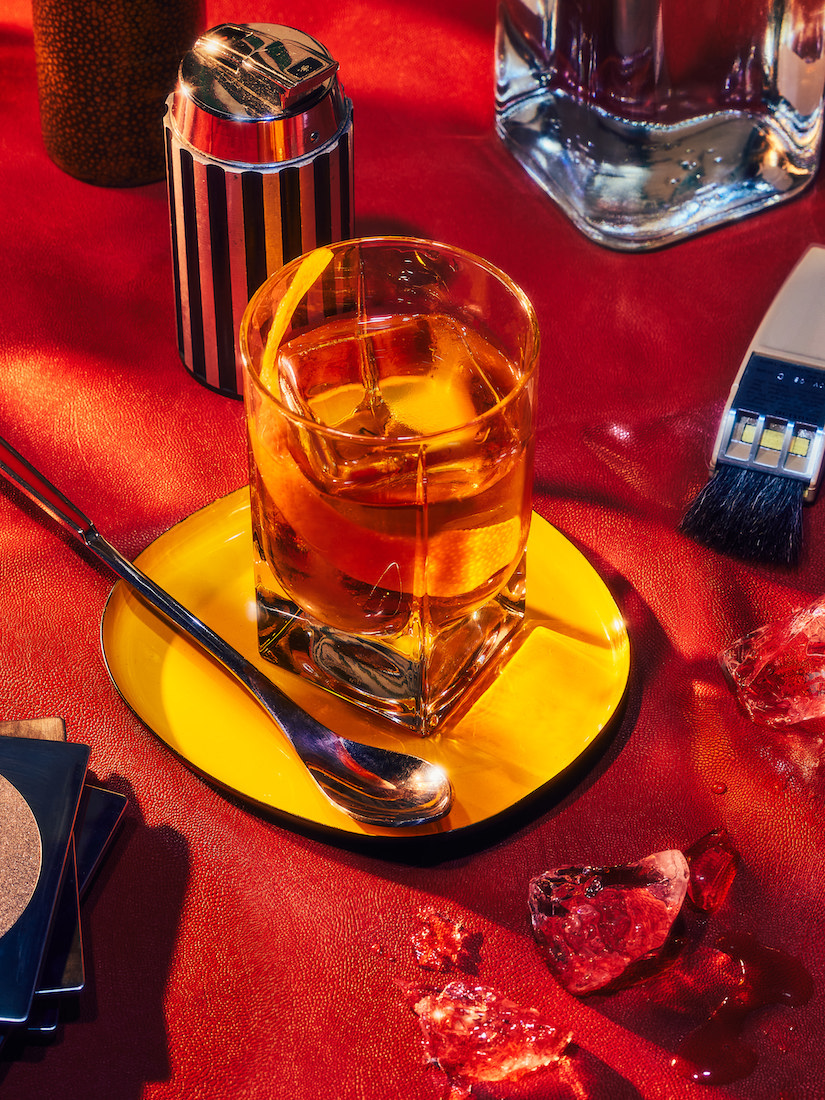 Crystal rocks glass containing a cocktail on a yellow saucer with a spoon, rock ice and art brush on a textured red surface.