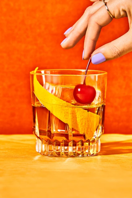 Close-up studio shot of a hand adding a cherry to an Old Fashioned cocktail with an orange peel, against an orange backdrop.