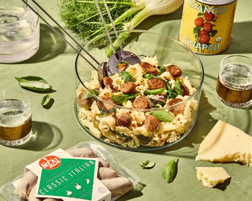 Overhead shot of Italian sausage pasta in a clear bowl, garnished with fresh basil, surrounded by fennel, beer, and ingredients.