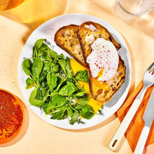 Overhead shot of a white plate with two slices of toast topped with poached eggs, a green salad, and a side of pepper.