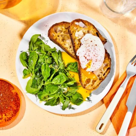 Overhead shot of a white plate with two slices of toast topped with poached eggs, a green salad, and a side of pepper.