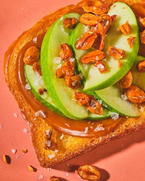 Toast topped with caramel spread, green apple slices, and caramelized nuts, presented on a pink surface with warm lighting.
