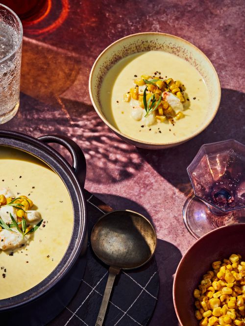 Overhead shot of creamy corn soup with charred corn, crab meat, and rosemary in bowls and a pot on a reddish-brown surface.