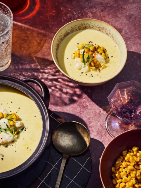 Overhead shot of creamy corn soup with charred corn, crab meat, and rosemary in bowls and a pot on a reddish-brown surface.
