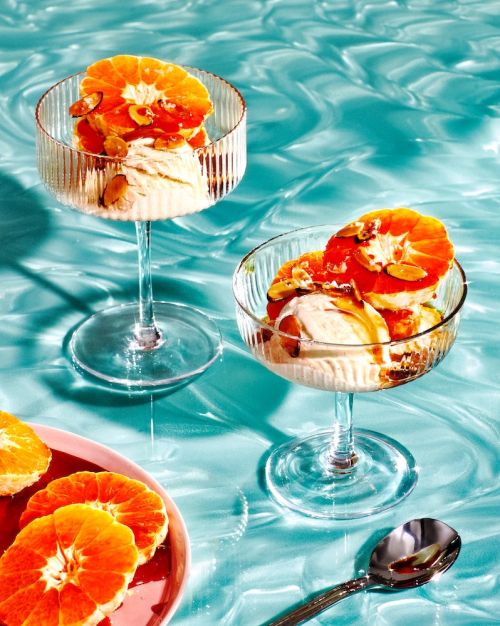 Two glass dessert cups filled with ice cream, garnished with orange slices and nuts, presented on a light blue, rippled surface.