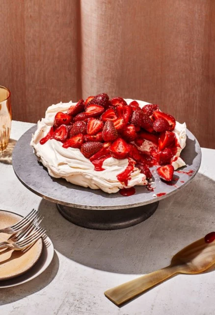 Overhead shot of a strawberry pavlova dessert on a grey stand, featuring meringue, cream, and fresh strawberries, with gold accents.