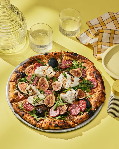 Overhead shot of a gourmet pizza topped with figs, sausage, and arugula on a metal pan, set against a bright yellow background.