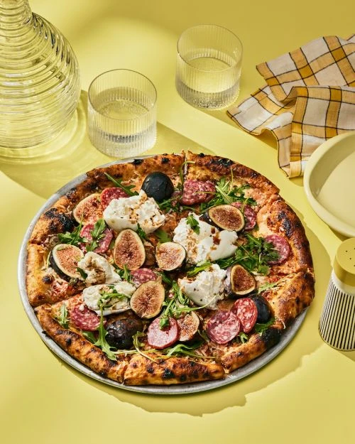 Overhead shot of a gourmet pizza topped with figs, sausage, and arugula on a metal pan, set against a bright yellow background.