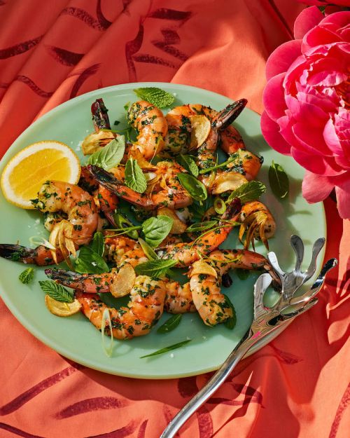 Overhead shot of grilled shrimp on a light green plate with lemon slices, herbs, and a coral patterned tablecloth; food photography.