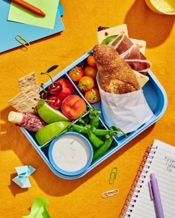 Overhead shot of a divided lunch box filled with a sandwich, cheese, fruit, vegetables, and crackers, along with school supplies on a yellow surface.
