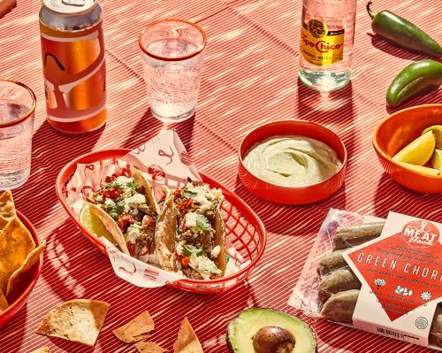 Overhead shot of tacos in red basket, surrounded by avocado, chips, drinks, limes, and chorizo on striped tablecloth.