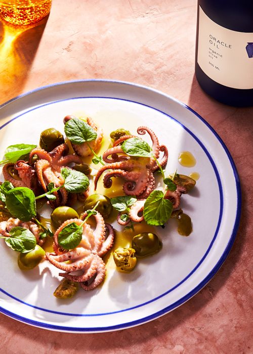 Overhead shot of an appetizer plate featuring octopus, green olives, and fresh greens, next to a bottle of olive oil.