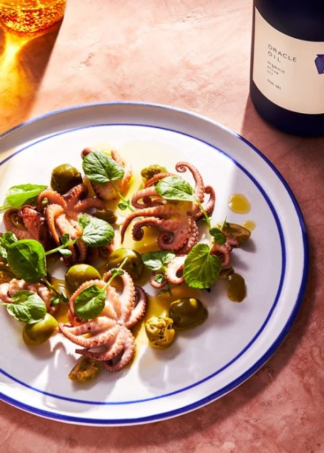 Overhead shot of an appetizer plate featuring octopus, green olives, and fresh greens, next to a bottle of olive oil.