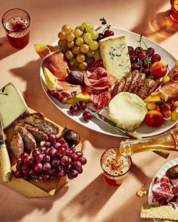 Overhead shot of a charcuterie board with various cheeses, meats, grapes, and a glass of beer being poured on a light peach surface.