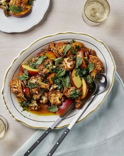 Overhead shot of a cauliflower and peach salad on a serving platter with fresh herbs and a side glass of white wine.