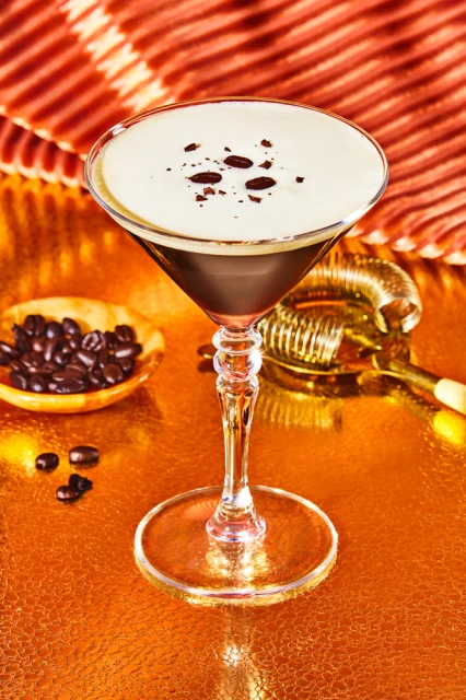 Espresso Martini cocktail in a stemmed glass with coffee beans on top and a metal cocktail strainer on an orange reflective surface.