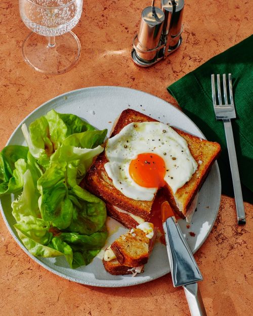 Overhead shot of egg-topped toast and lettuce salad on a plate, next to a glass of water and salt and pepper shakers.