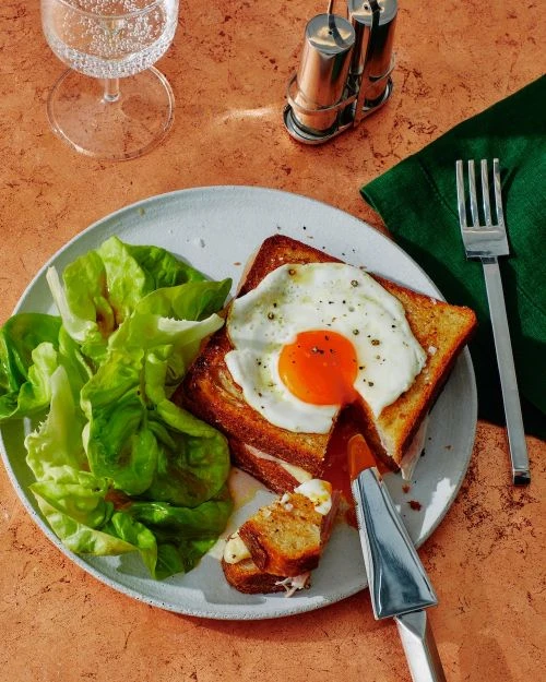 Overhead shot of egg-topped toast and lettuce salad on a plate, next to a glass of water and salt and pepper shakers.
